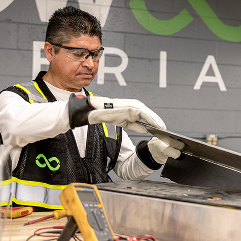 Photograph of a Redwood Materials technician wearing safety glasses, gloves, and a black logoed vest with reflective stripes working with a metal sheet in an industrial setting. The background features a gray wall with partially visible green and white text, and a yellow multimeter is placed on the workbench in the foreground.