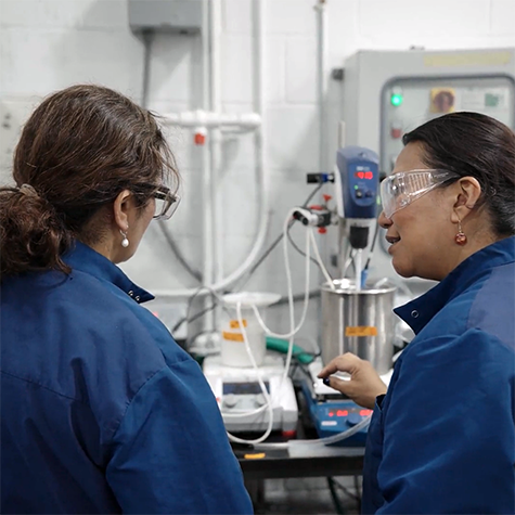 Photograph of two female employees in blue lab coats and safety glasses discussing equipment in a laboratory setting. The background includes various instruments, wires, and control panels, indicating an active experimental or research environment.