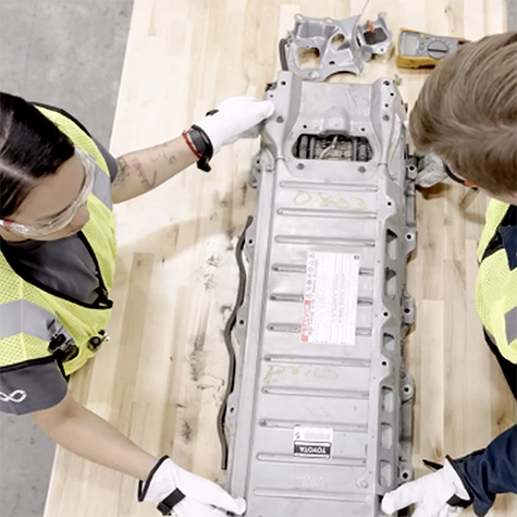 Photograph showing two workers wearing safety vests and gloves examining a large metallic automotive component labeled "Toyota" on a wooden table. The scene highlights a collaborative inspection or assembly process in an industrial or manufacturing setting.