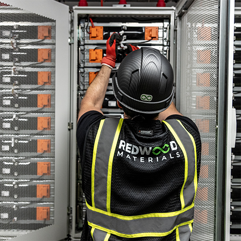 Photograph of a technician wearing a black helmet and a high-visibility Redwood Materials logoed vest working on a battery storage unit with orange connectors inside a metal cabinet. The scene highlights maintenance or assembly of energy storage components, emphasizing safety gear and organized battery modules.